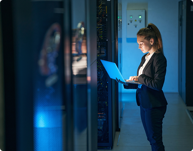 a professionally dressed woman holding a laptop standing in a server room